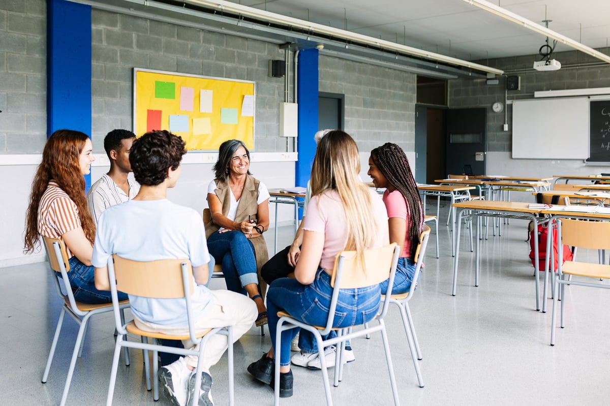 Young group of teenage students discussing in classroom sitting in circle with female teacher